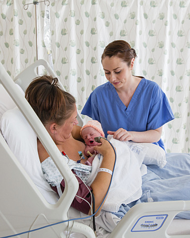 Mum in bed with new baby being attended to by healthcare worker.